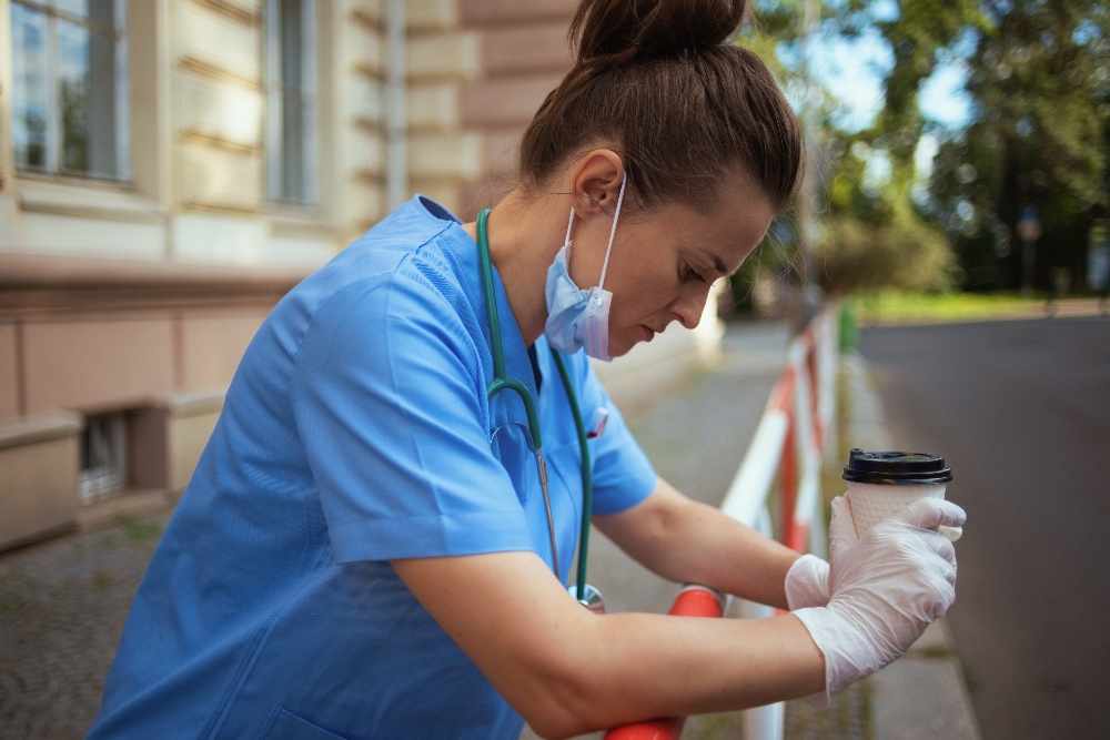 A female nurse in scrubs, surgical mask, and protective gloves stands outside of a hospital with a coffee cup, looking down. She appears to be stressed.