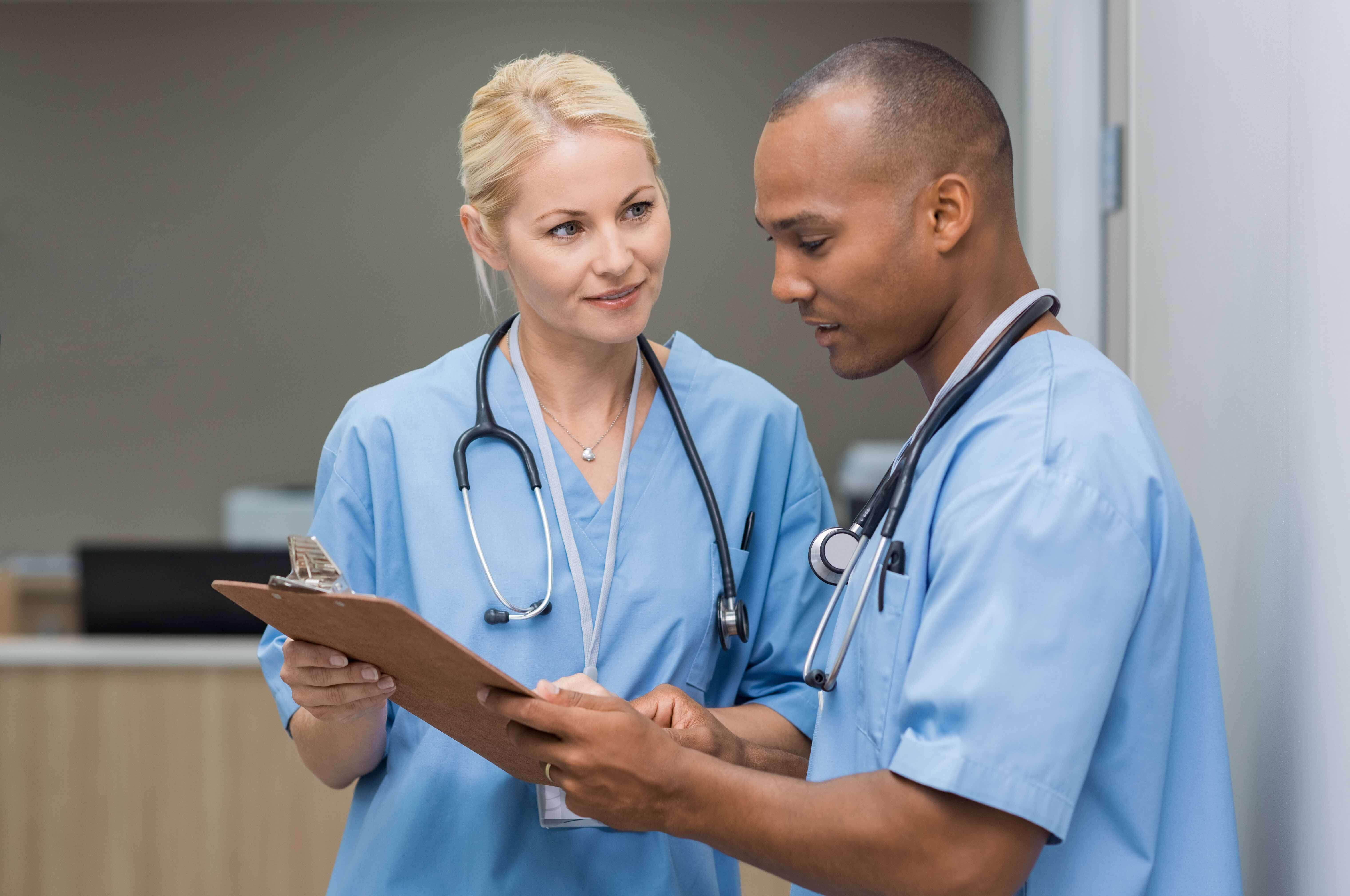 Female nurse leader in blue scrubs holds clipboard with male nurse in blue scrubs, teaching him how to properly log medical records.
